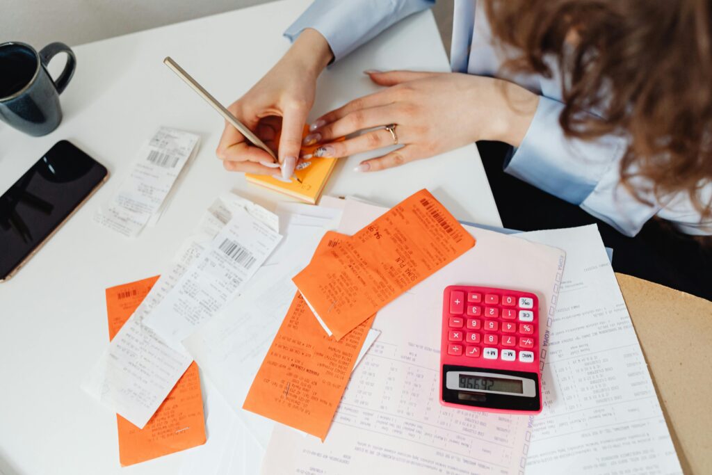 pexels-photo-7680751-7680751 A woman writes down financial details using a calculator, receipts, and documents.